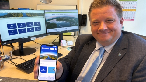 Bürgermeister, der neue Homepage auf dem Handy zeigt, im Hintergrund sind zwei Bildschirme