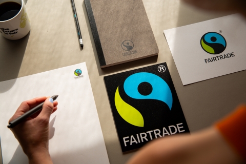 Block, Stift und Fairtrade-Zeichen (blau-grüner Kreis mit stilisierter Figur)