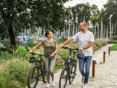 Eine Frau und ein Mann, die ihr Fahrrad am See langschieben.
