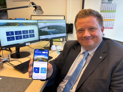 Bürgermeister, der neue Homepage auf dem Handy zeigt, im Hintergrund sind zwei Bildschirme